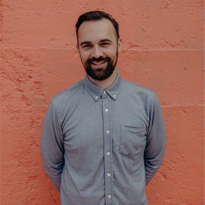 Smiling person in a gray button-up shirt standing against a textured orange wall.